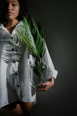 A woman in a white shirt holds a fern leaf in a glass vase, fashioned artfully indoors.